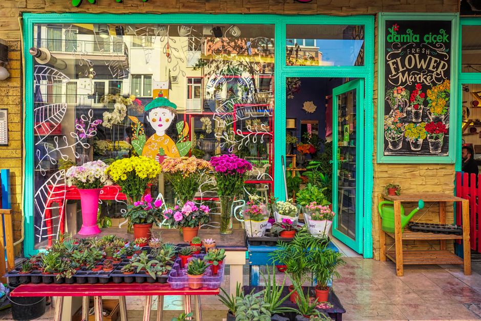 The flower shop features a vibrant turquoise door and window frame, showcasing an array of fresh potted flowers and floral arrangements on display. Brightly colored chrysanthemums in pink, yellow, whi