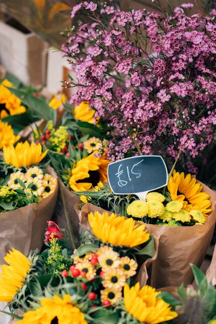 A vibrant display of fresh flower bouquets featuring yellow sunflowers, small white daisies, and red accents, wrapped in brown kraft paper. Behind the bouquets, a large cluster of pink and purple waxf